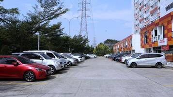 Un parking où de nombreuses voitures sont garées en rangées, un grand immeuble à la façade à motifs en damier sur la droite, et des arbres en arrière-plan.