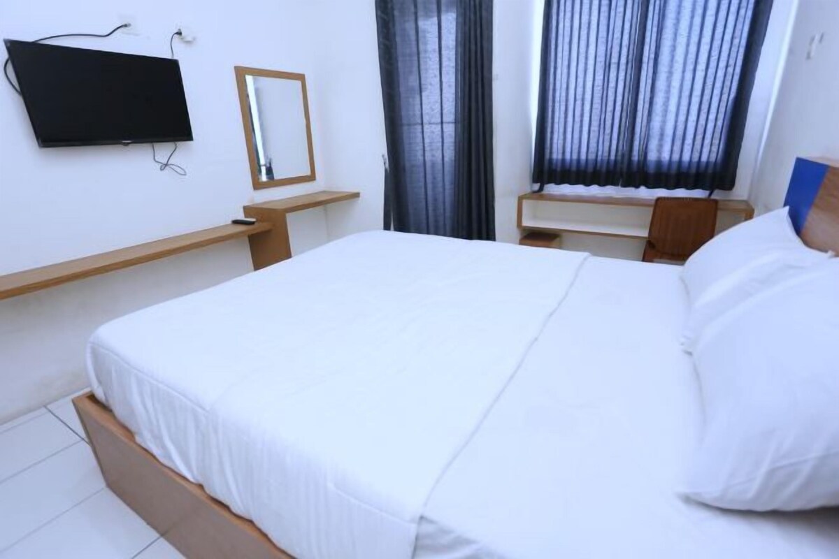 Ett hotellrum med en säng, en tv som hänger på väggen, en spegel och ett fönster med gardiner.