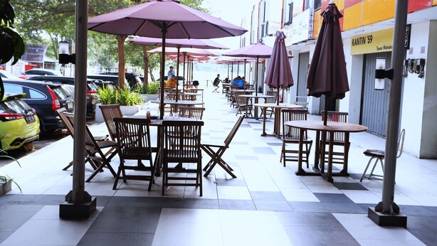 An outdoor cafe with tables and chairs, umbrellas, and a building with a sign in the background. - Nusantara Rooms Mahakam By Reccoma (Cikarang)