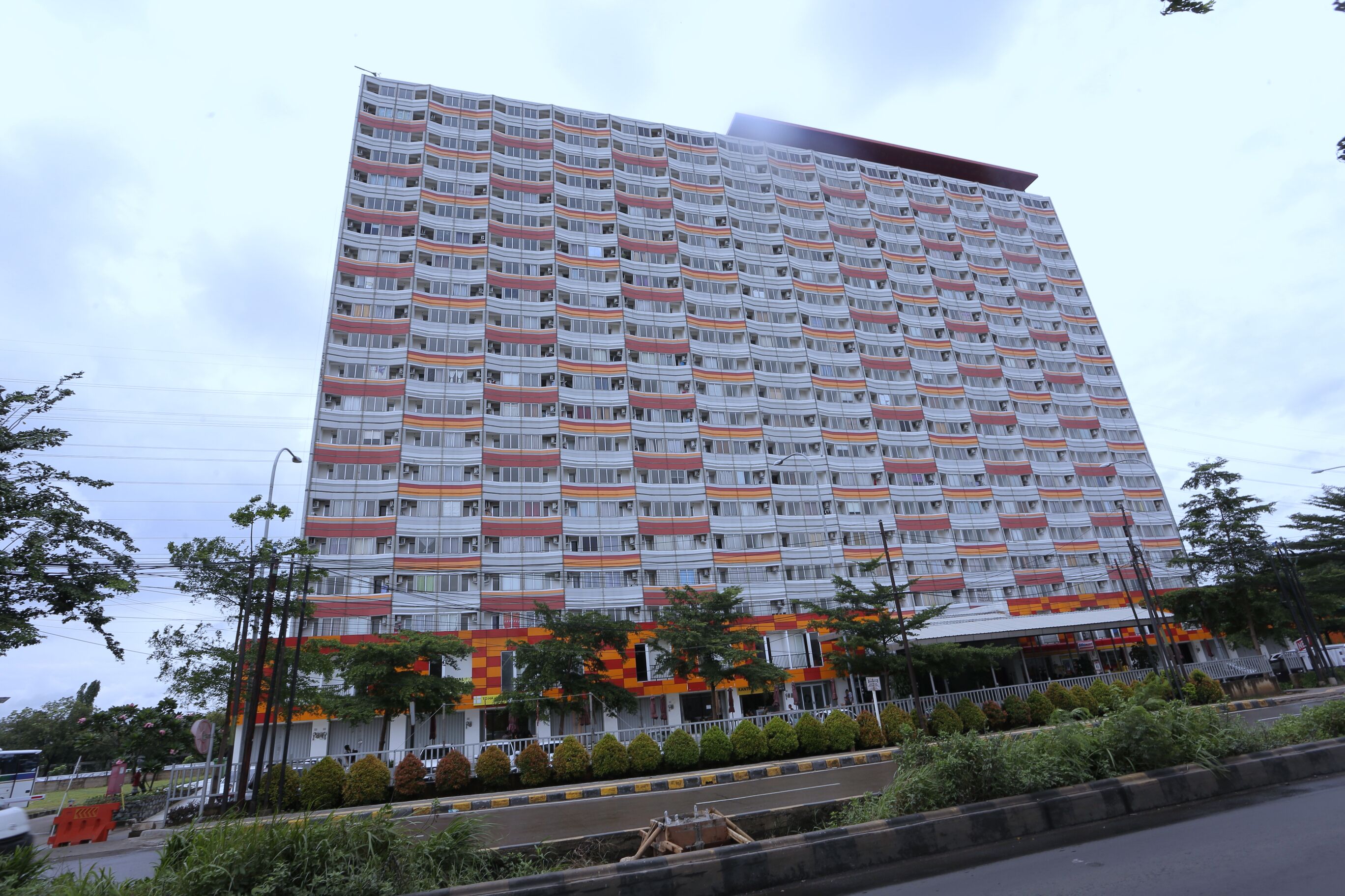A multi-story building with balconies, a road in front, and some greenery.