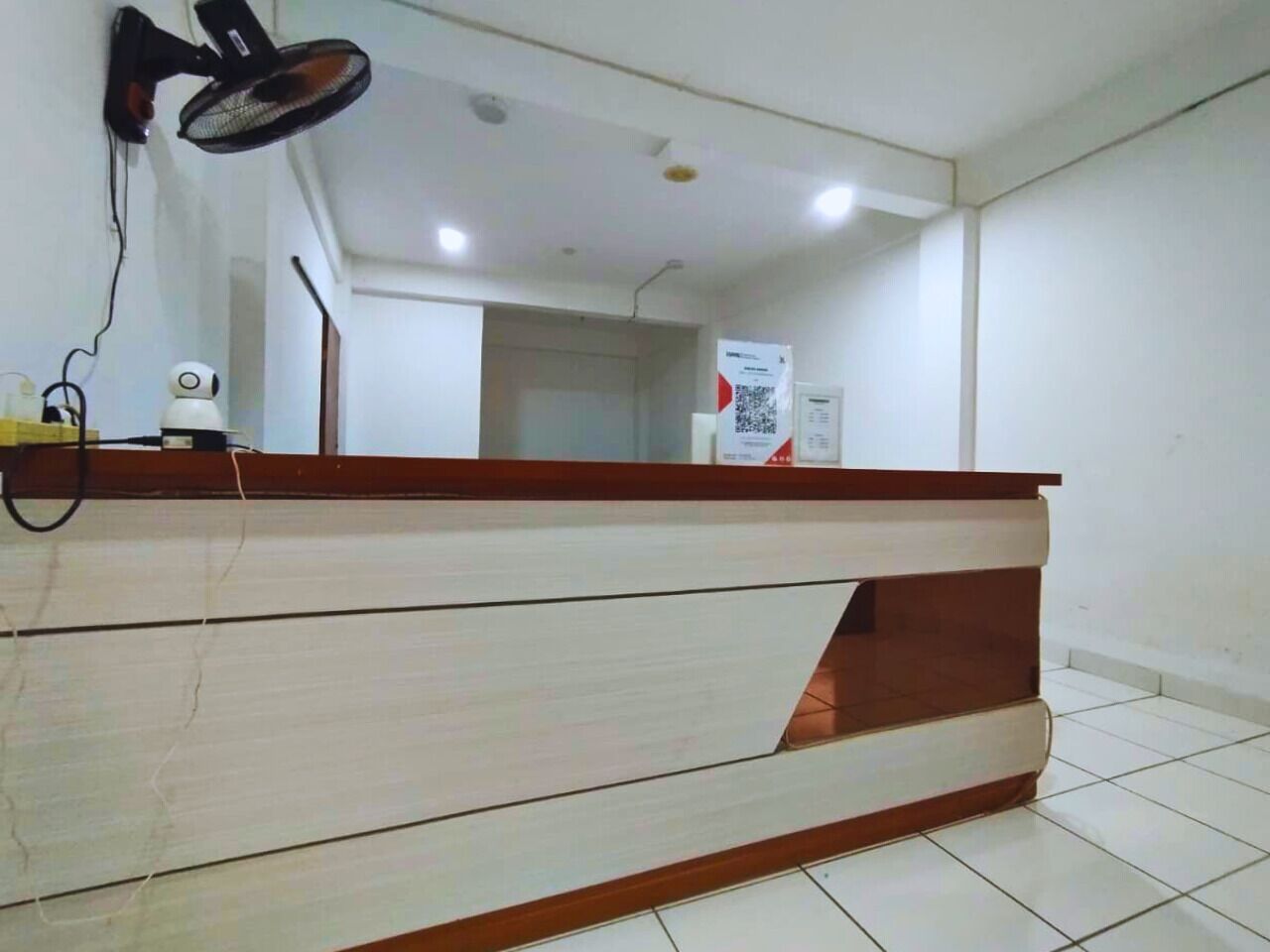 A reception desk with a wooden top and a light-colored panel, a wall-mounted fan, and a poster on the wall.