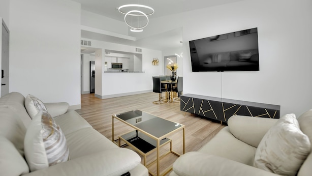 A modern living room with a sofa, a glass coffee table, a TV mounted on the wall, and a dining area in the background. - Charming 2-bedroom condo near to Strip (Las Vegas)