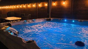 Un bain à remous avec un mur en bois et des guirlandes lumineuses, entouré de fauteuils en osier et d’une table.