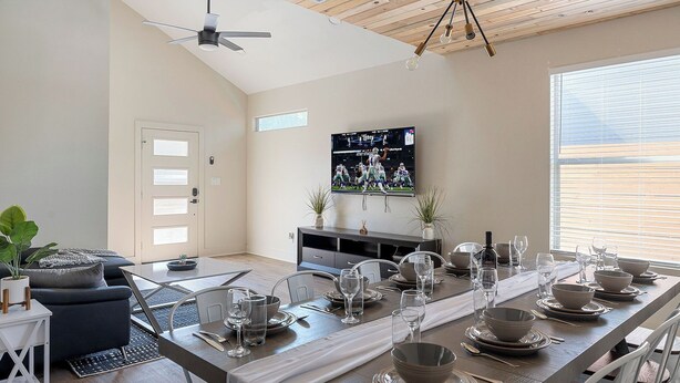 Un espace repas moderne avec une longue table dressée pour six personnes, un ventilateur de plafond et un téléviseur fixé au mur.