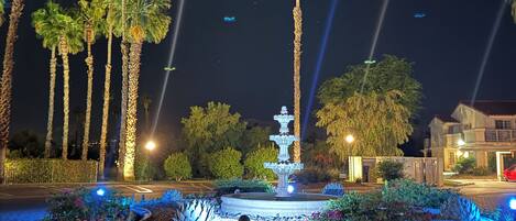 A nighttime view of a landscaped area with a central fountain, palm trees, and a paved walkway.