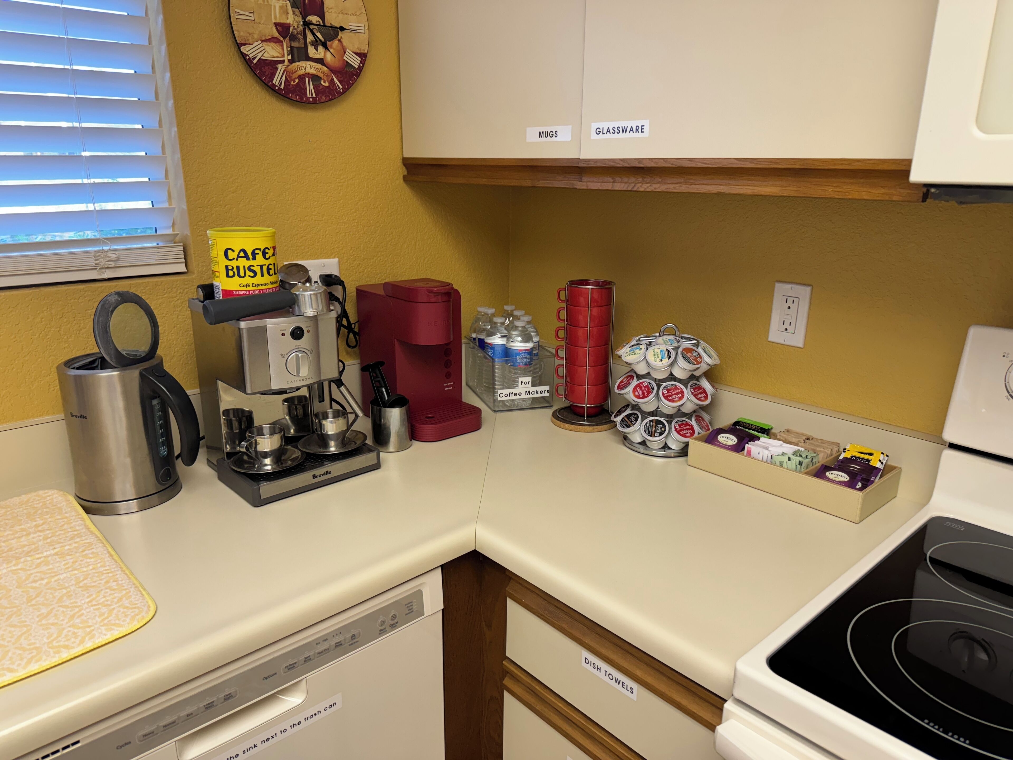 Cozinha equipada com cafeteira, chaleira, espaço para café e armários identificados para canecas e copos.