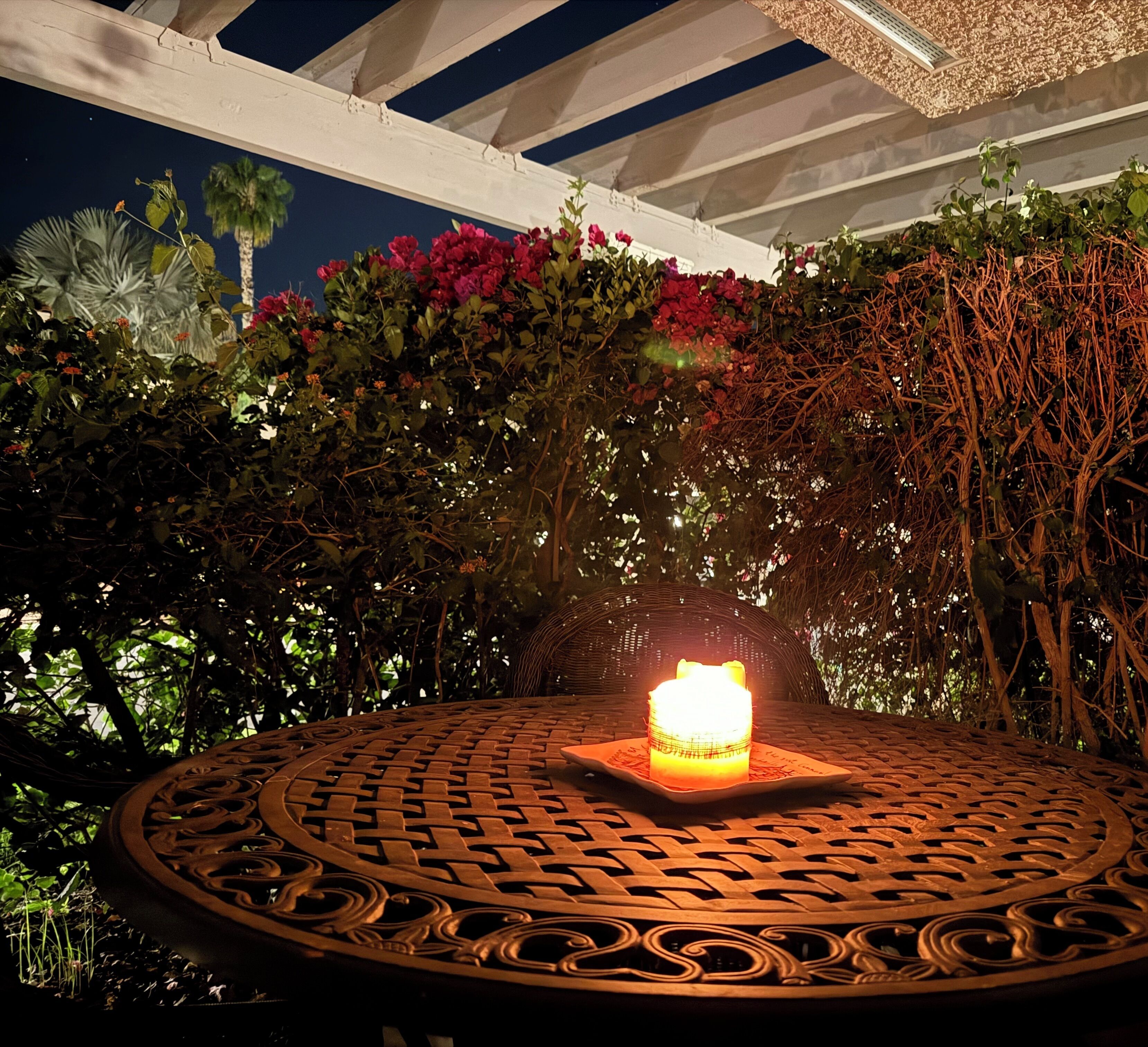 Une table ronde placée à l’extérieur, avec une bougie au centre, entourée d’une végétation luxuriante et d’une structure de pergola blanche.