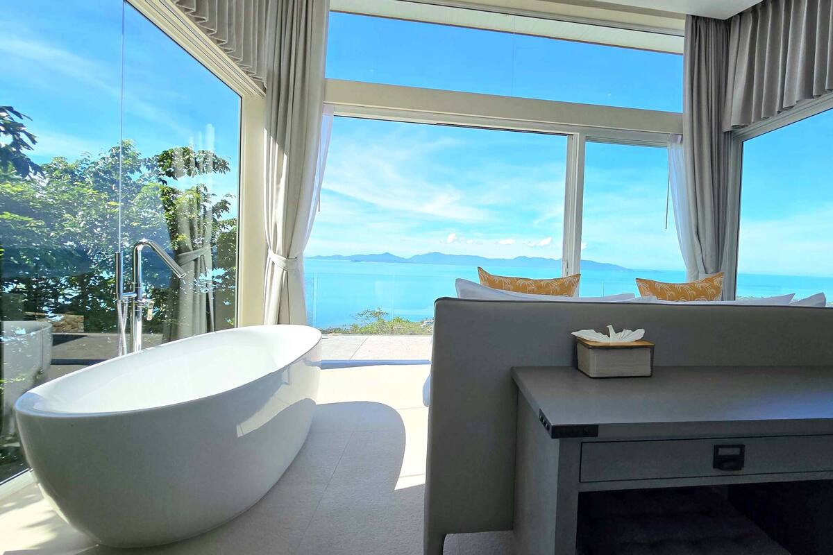 A modern bathroom with a freestanding bathtub, a vanity with a mirror, and a view of the sea.