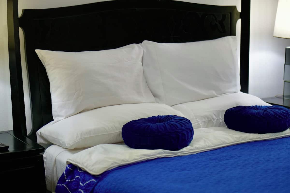 Een hemelbed met witte lakens en twee blauwe kussens, een nachtkastje met een lamp, en een blauwe sprei.