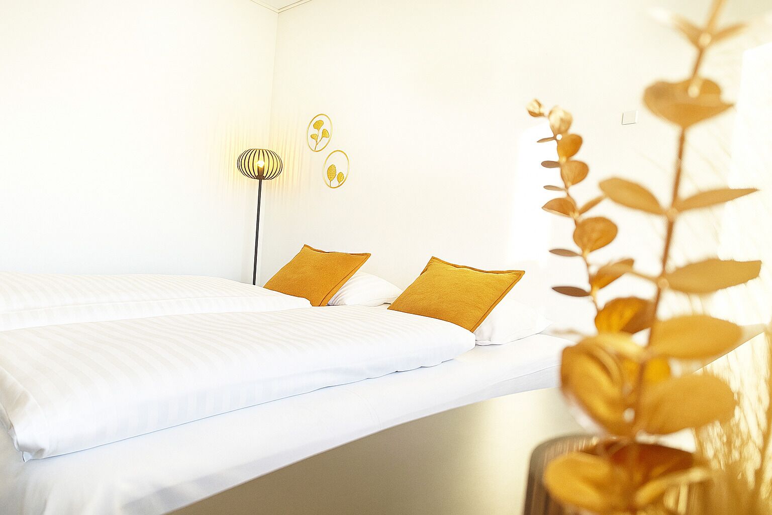 Hotellihuone, jossa on sänky, kaksi tyynyä, lattiavalaisin ja seinätaideteos.