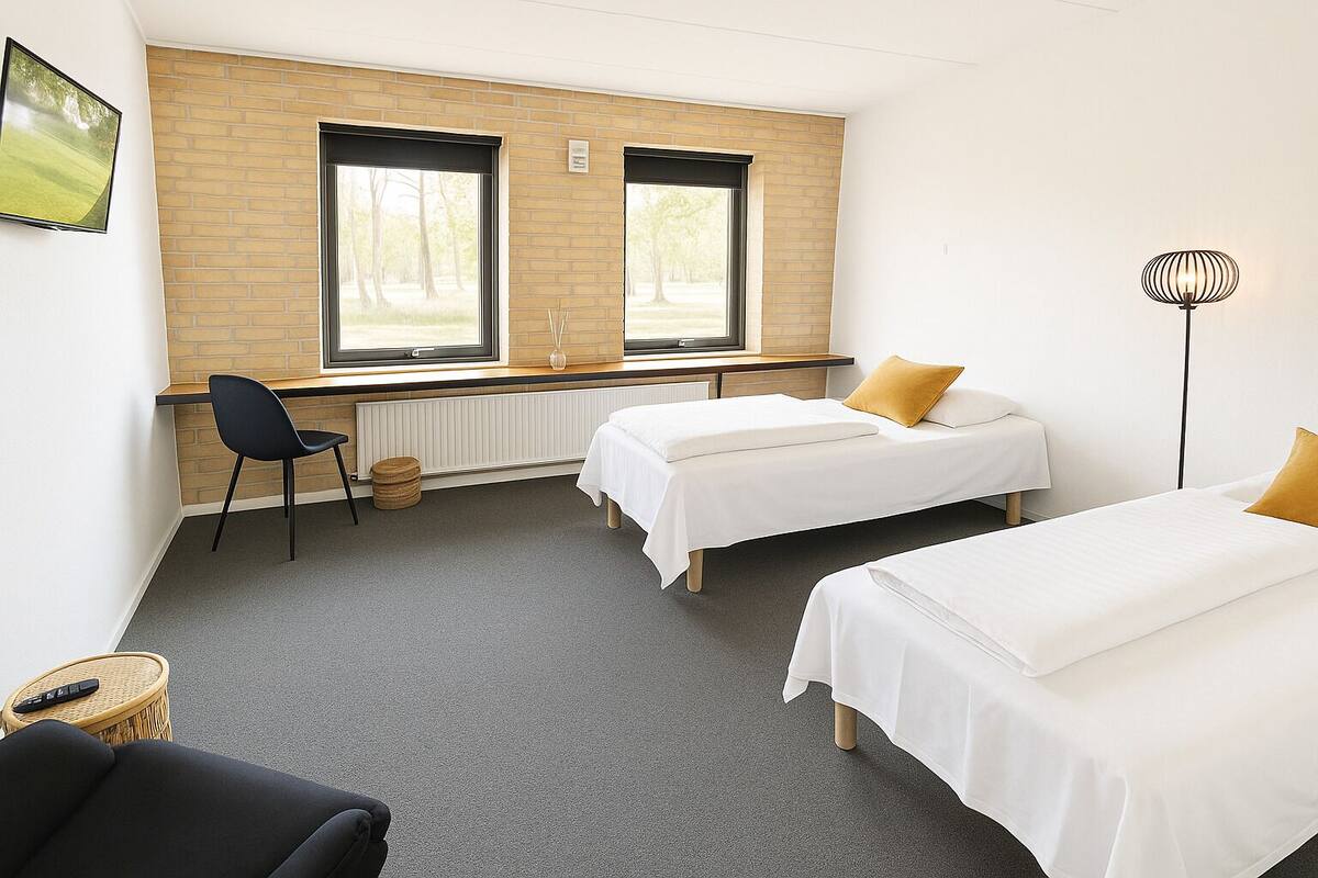 A room with two beds, a desk, a chair, a lamp, and a view of trees through large windows.