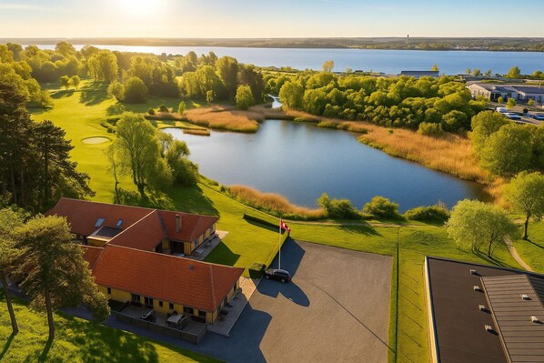 A golf course with a lake, surrounded by trees and a building with a red roof. - Hotel Svanegården (Korsor)