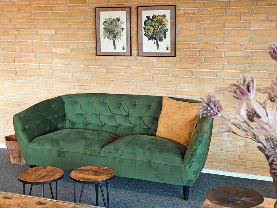 Ein grünes Sofa mit braunem Kissen, zwei kleine runde Tische und zwei gerahmte Pflanzenbilder an einer Backsteinwand.