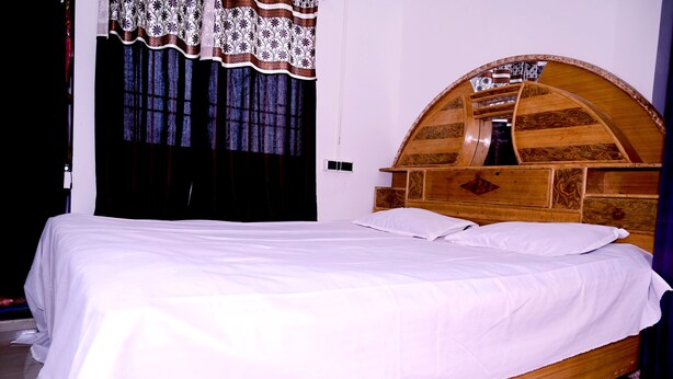 A single bed with a wooden headboard, white bedding, and a window with patterned curtains. - SHIVAY GUEST HOUSE (Deoghar)