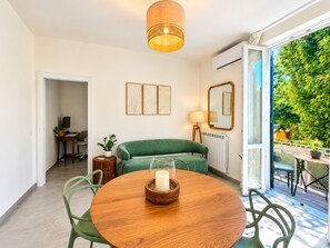 A modern living room with a round wooden table, green chairs, a green sofa, a hanging lamp, and a view of a green garden through a glass door.