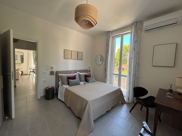 Een slaapkamer met een bed, een bureau en een stoel. Er is een raam met gordijnen, een deur en een wandgemonteerde airconditioning.