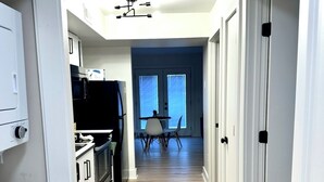 A narrow hallway with a kitchen on one side, a dining area with a table and chairs, and a door leading to another room.