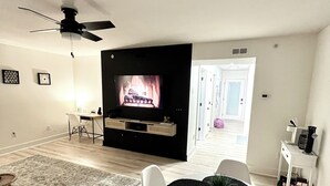 A modern living room with a flat-screen TV mounted on a black wall, a ceiling fan, a dining table with white chairs, and a large rug on the floor.