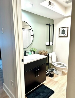 Une salle de bain avec un miroir rond, un lavabo blanc, un meuble noir, des toilettes et un tableau encadré au mur.