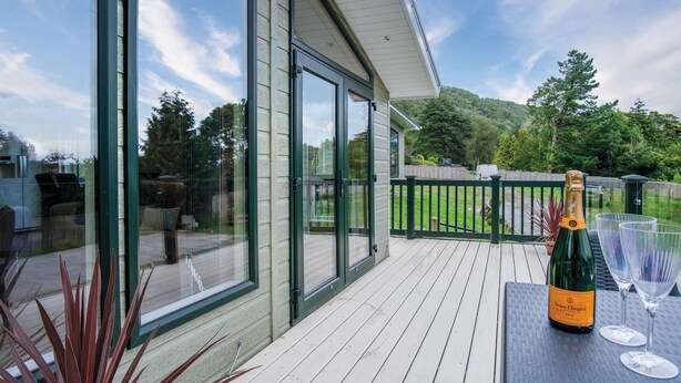 A balcony with a bottle of champagne and glasses, a wooden deck, and a glass door leading to a patio area. - 3 Manor Lodge - 2 Bedroom Lodge - Pendine (Carmarthen)