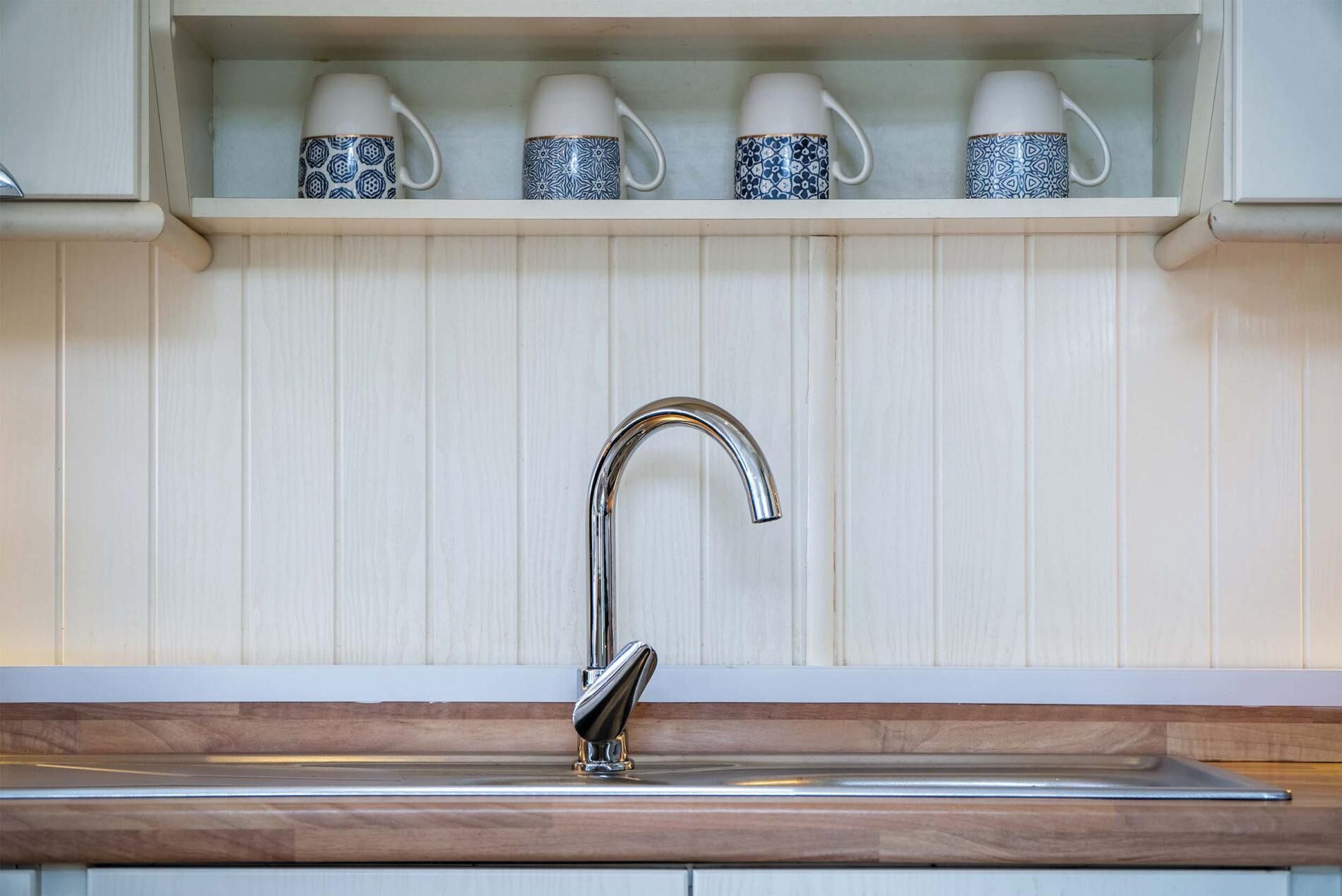 A kitchen with a stainless steel sink, a white backsplash, and a shelf above holding four patterned cups.