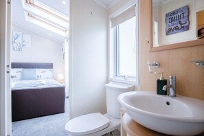 A modern bathroom with a white sink, a toilet, and a shower. There is a window with a towel hanging on the door. - 2 Manor Lodge - 2 Bedroom Lodge - Pendine (Carmarthen)