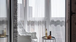 Une chambre moderne avec une porte vitrée donnant sur un balcon, un fauteuil blanc, une petite table avec une bouteille et des raisins, et une vue sur la ville.