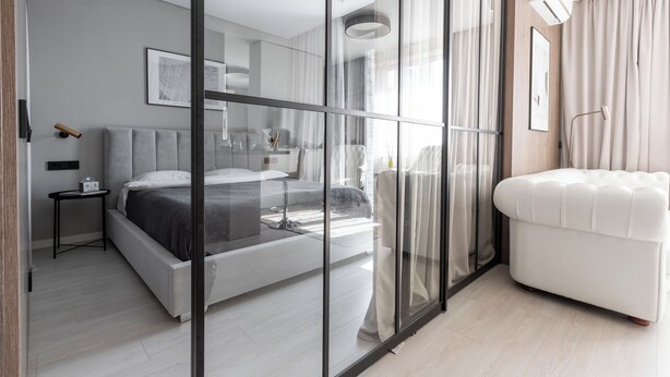 Une chambre moderne avec un grand lit, une table de chevet, une commode, un canapé blanc et une grande fenêtre ornée de rideaux légers.