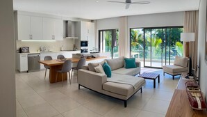 Een moderne woonkamer met een bank, salontafel en eethoek. Achtergrond is een keuken met witte kastjes en een eettafel.