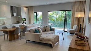 Een moderne woonkamer met een bank, eettafel en stoelen. Er is een keukenhoek met kasten en een raam met uitzicht op een tuin.