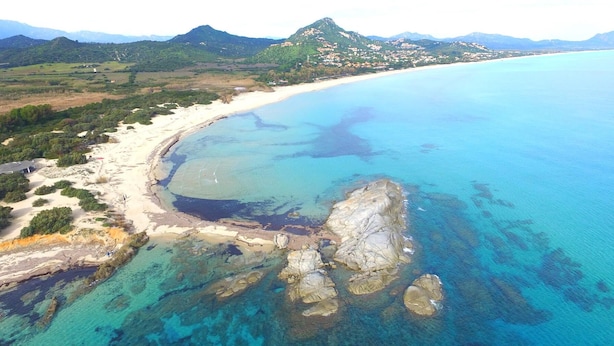 Vue aérienne d’une zone côtière avec une plage de sable, des eaux bleues et limpides, et des affleurements rocheux.