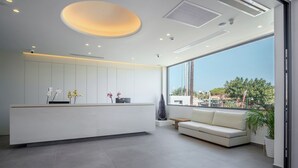 Un espace d’accueil moderne, doté d’un comptoir de réception blanc, d’un canapé et d’une grande fenêtre donnant sur l’extérieur.