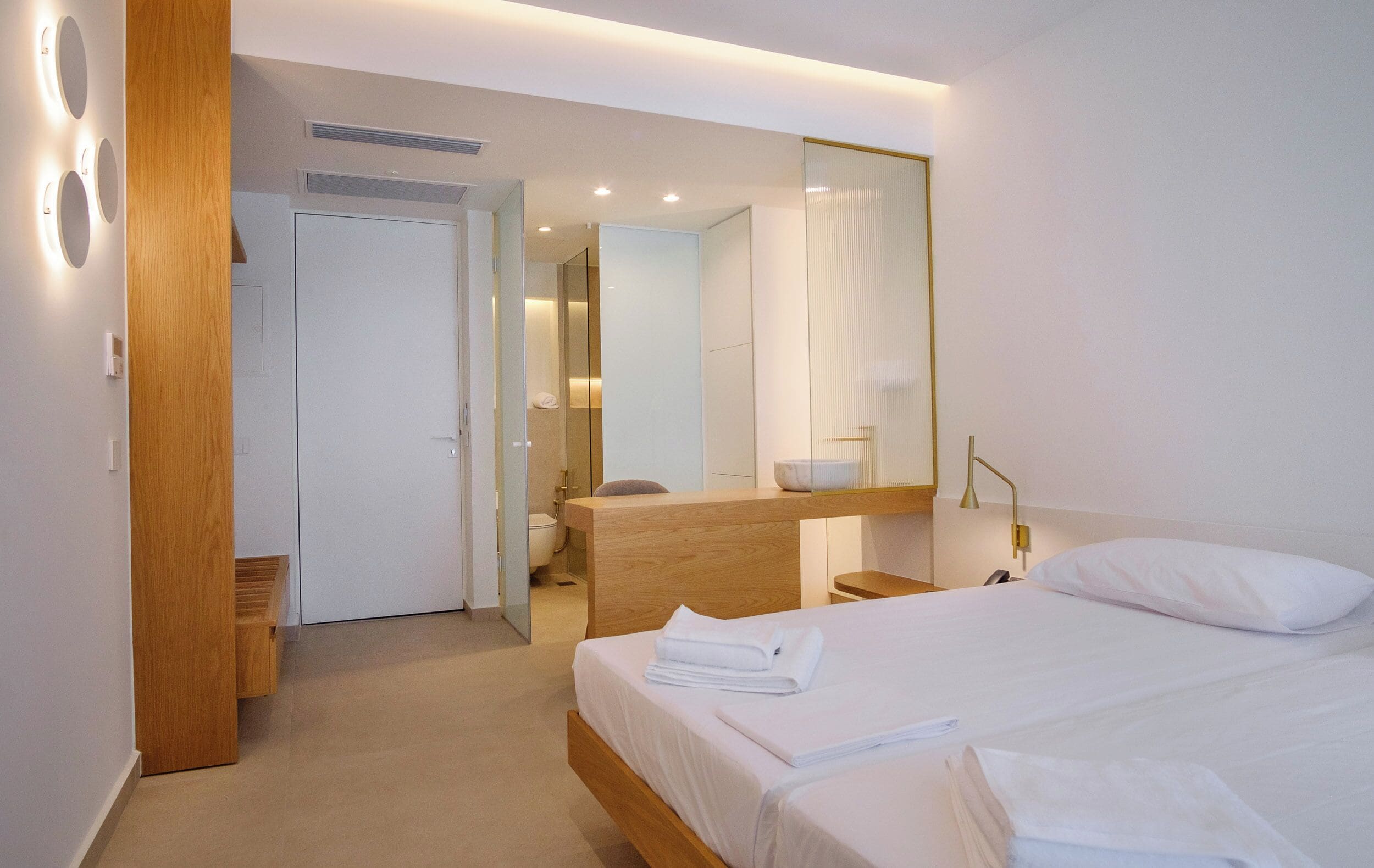A modern hotel room with a large bed, a wooden nightstand, a mirror, and a bathroom visible through an open door.