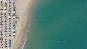 Vue aérienne d’une plage avec des rangées de parasols et de chaises de plage, un rivage sablonneux et une eau d’un bleu limpide.