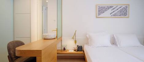 A hotel room with a wooden desk, a bed with white linens, a bedside table with a lamp, and a framed abstract artwork on the wall.