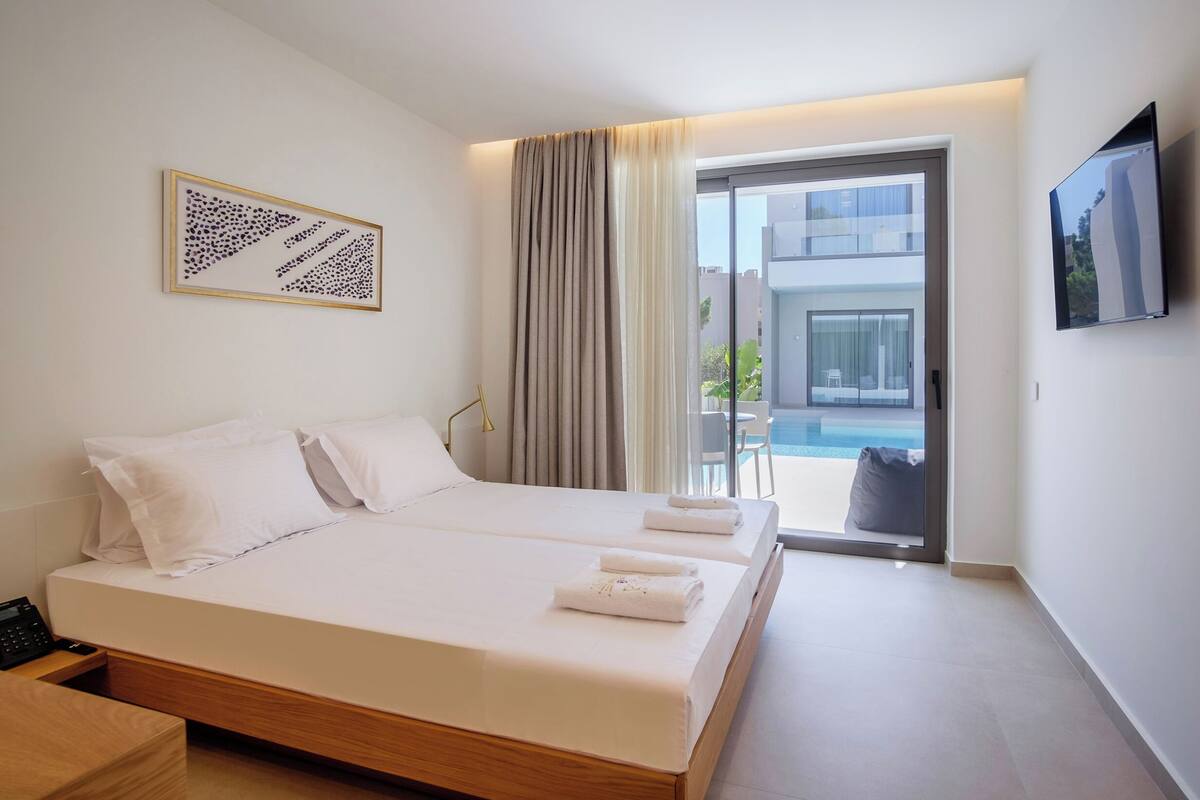 Ett modernt hotellrum med en stor säng, ett sängbord, en platt-TV och utsikt över en pool genom en skjutdörr i glas.