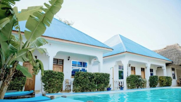 A poolside view of a white building with blue roofing, surrounded by lush greenery and a clear blue pool. - Serenity Boutique Hotel (Jambiani)