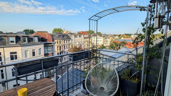 Un balcón con vistas a un paisaje urbano, un ventilador grande y una taza amarilla.