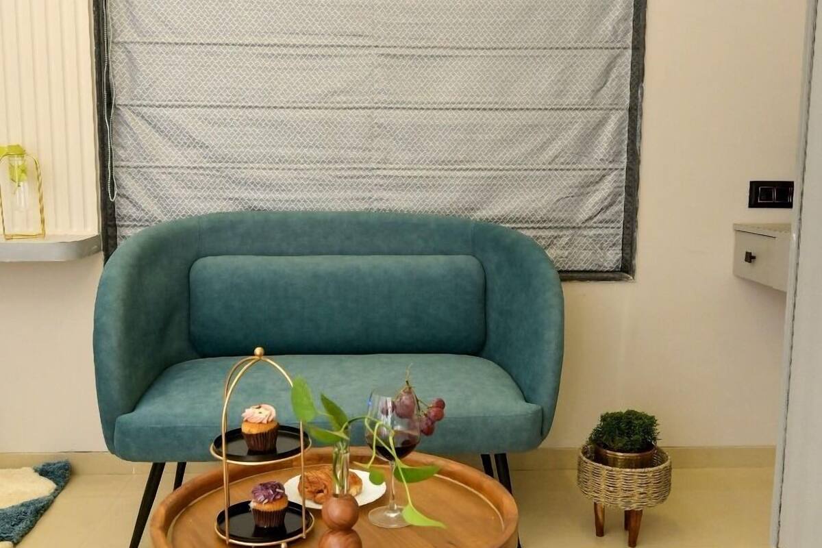 A teal sofa against a white wall with a window blind, a wooden coffee table with a cupcake and a vase, and a small potted plant.