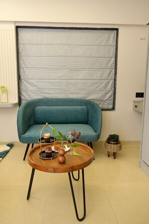 Un canapé de couleur turquoise est placé contre un mur blanc, près d’un store de fenêtre. On y voit également une table basse en bois sur laquelle sont posés un cupcake et un vase, ainsi qu’une petite plante en pot.
