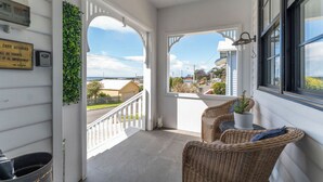 A covered porch with wicker furniture and a potted plant, overlooking a residential street. - Ocean Breeze - Modern Comfort with Outdoor Entertaining and Ocean Views (Parklands)