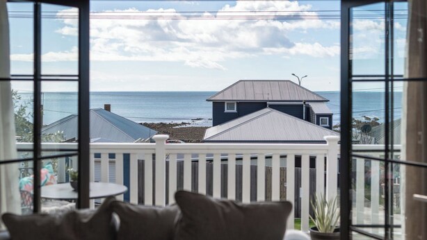A room with a view of the sea through large windows, a white fence, and a small table. - Ocean Breeze - Modern Comfort with Outdoor Entertaining and Ocean Views (Parklands)
