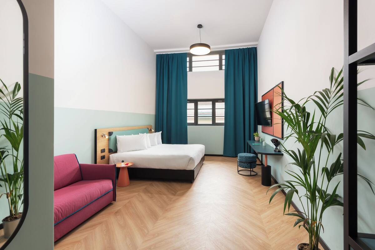 A modern hotel room with a bed, a pink sofa, a small table, and a potted plant.