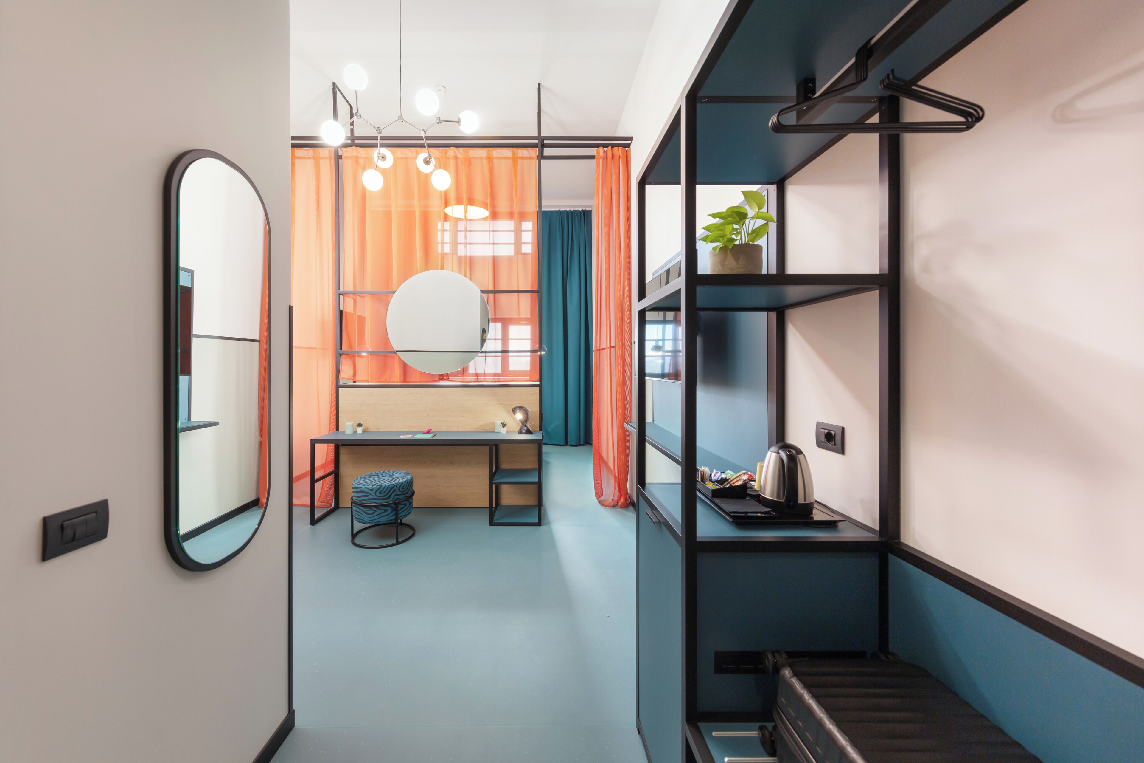 A compact room with a mirror, a desk with a chair, a hanging light fixture, and a shelf with a plant.