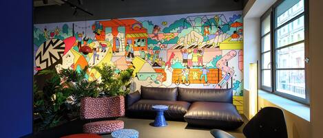 A modern lounge area with a colorful mural, comfortable seating, and a large window providing a view of the city.