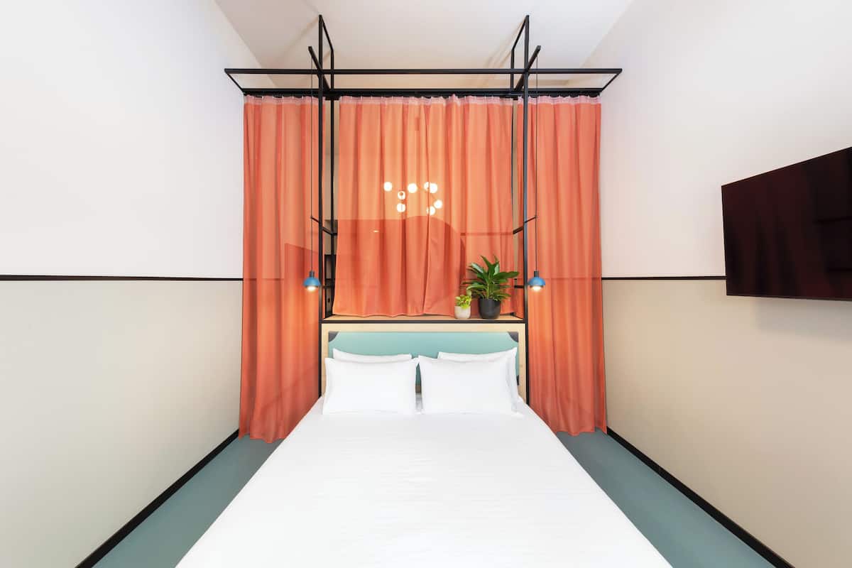 A modern hotel room with a bed, orange curtains, a potted plant, and a mounted TV.