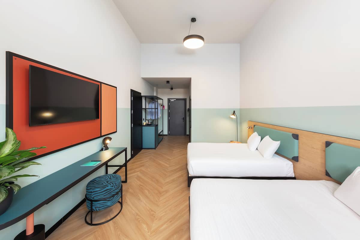 A hotel room with a large TV, a desk with a chair, two beds, and a potted plant.