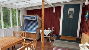 Un porche cubierto con una mesa de madera, una silla a rayas, una mesita con un farol y una puerta con una bandera.