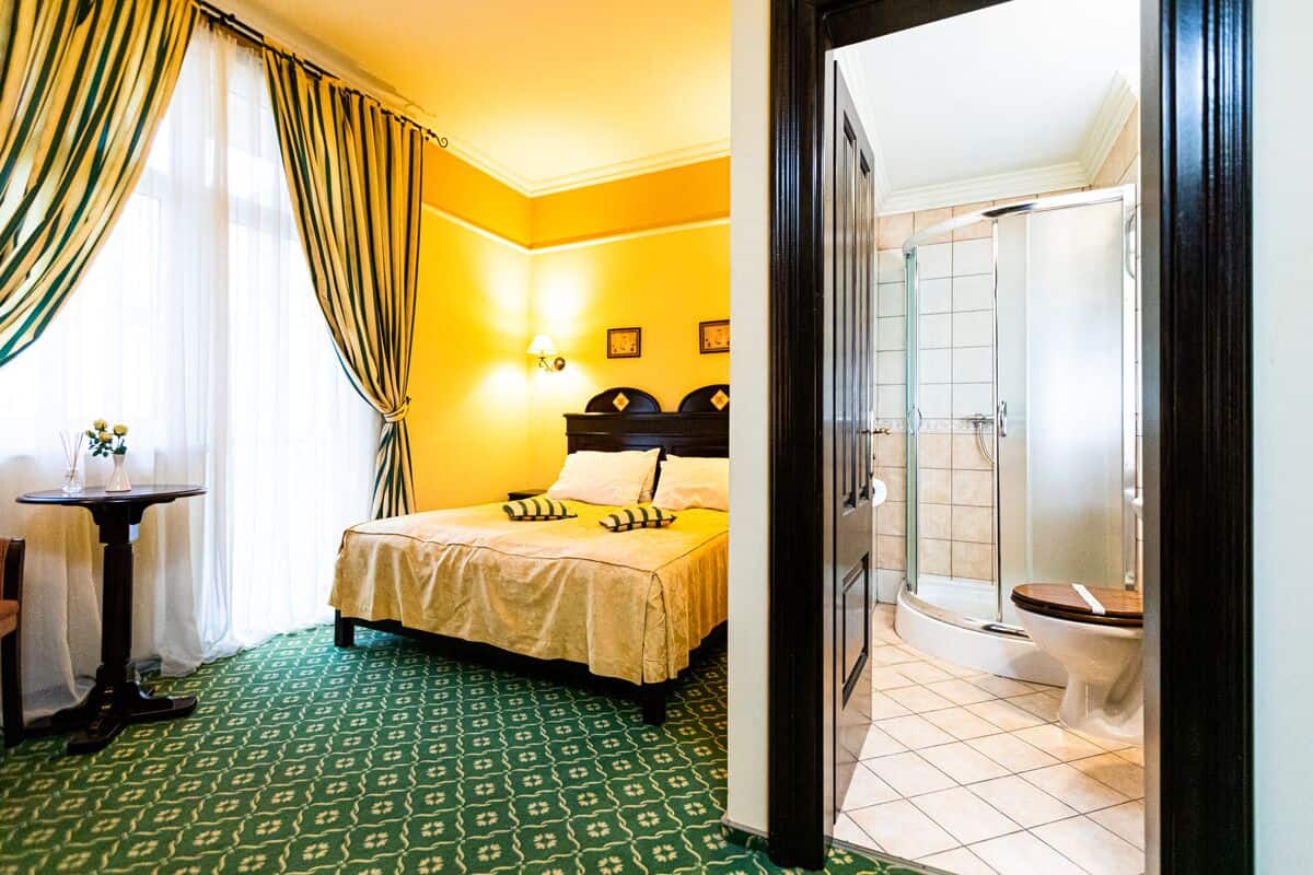 A hotel room with a bed, a small table with a vase, a lamp, and a shower area.