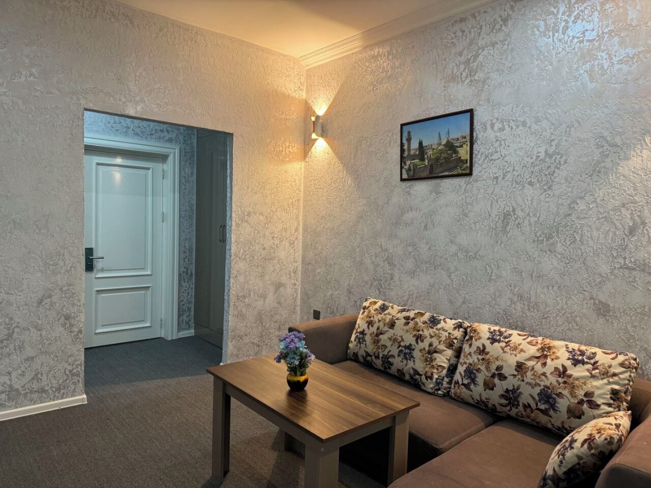 Ett vardagsrum med en soffa, ett träbord med en vas med blommor, en vägglampa och en inramad tavla.